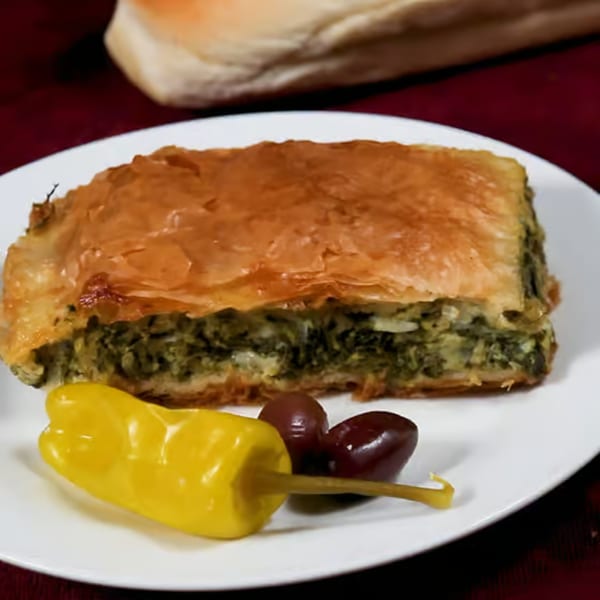 A slice of Greek spanakopita with a yellow pepper and two olives on a white plate.