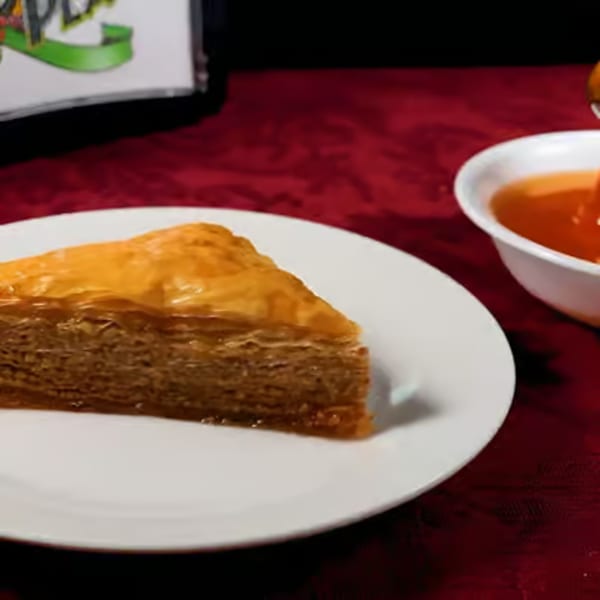 A slice of Greek baklava with a side of honey on a white plate and red tablecloth.